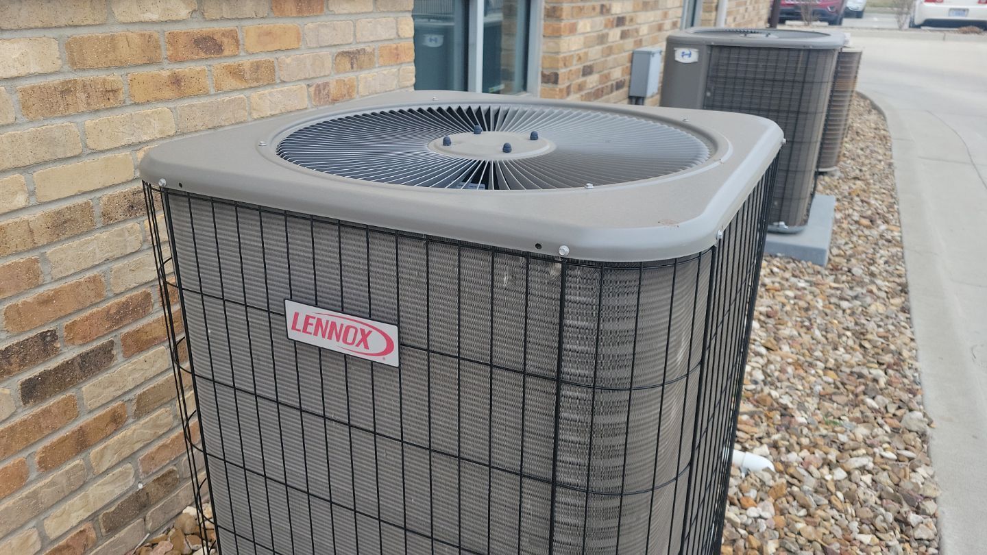 Lennox air conditioning unit outside a brick building, near a road, with a gray metal mesh exterior.