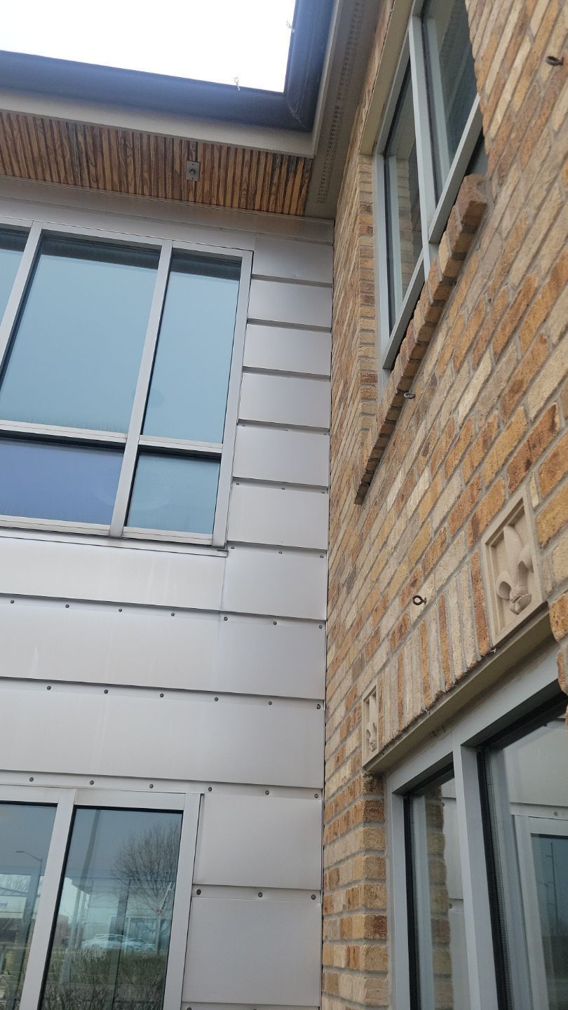 Exterior building corner: brick wall joins metallic siding, windows on both sides, overcast sky.