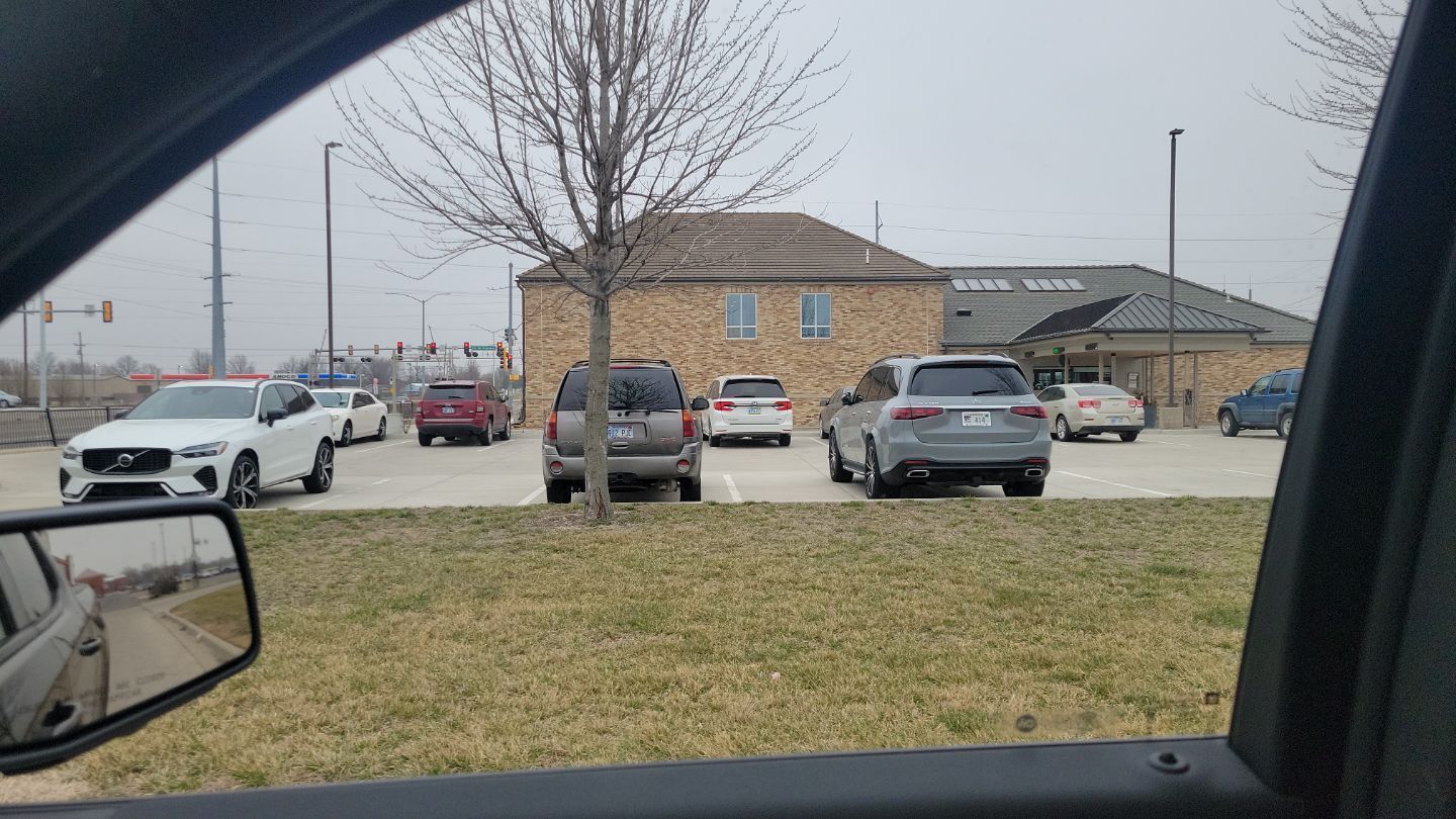 View from a car window of a parking lot in front of a brick building on a cloudy day. Several cars are parked.