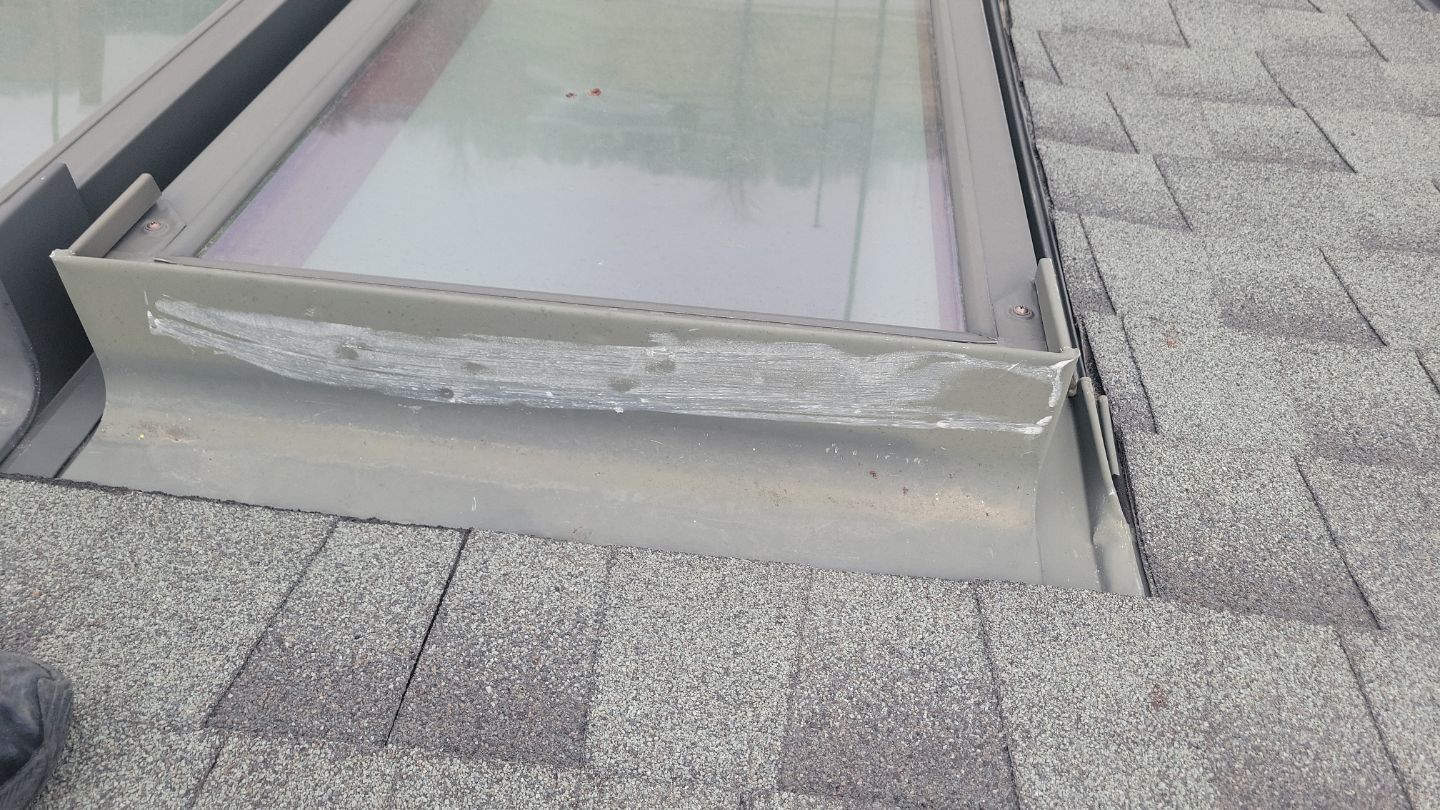 Skylight flashing with weathered sealant on a shingled roof, showing potential leak points.