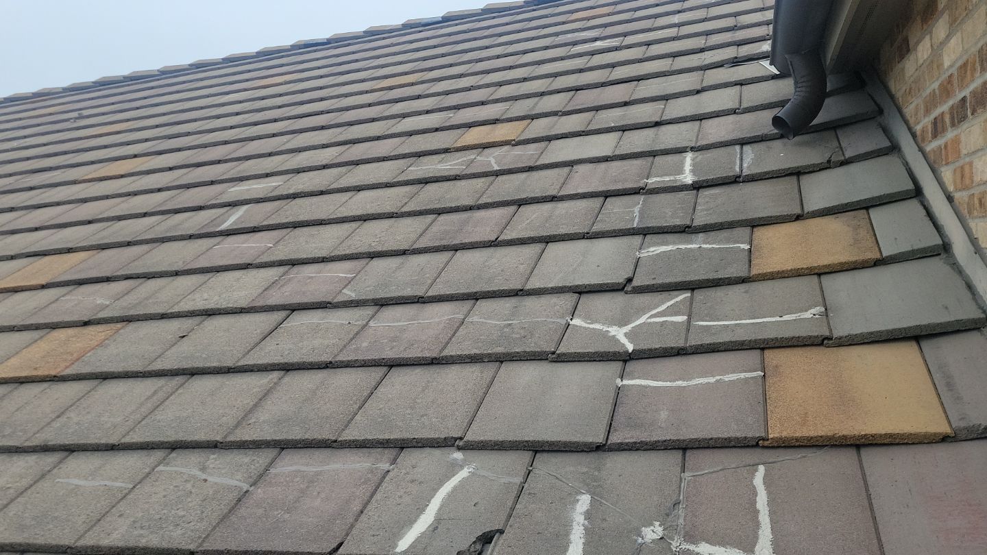 Close-up of a roof with multiple cracked tiles, some marked with white sealant; next to a brick wall.