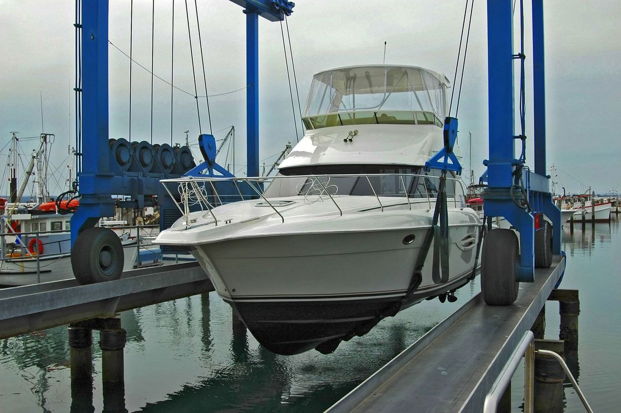 A white boat being lifted out of the water by a blue marine travel lift. Harbor background.