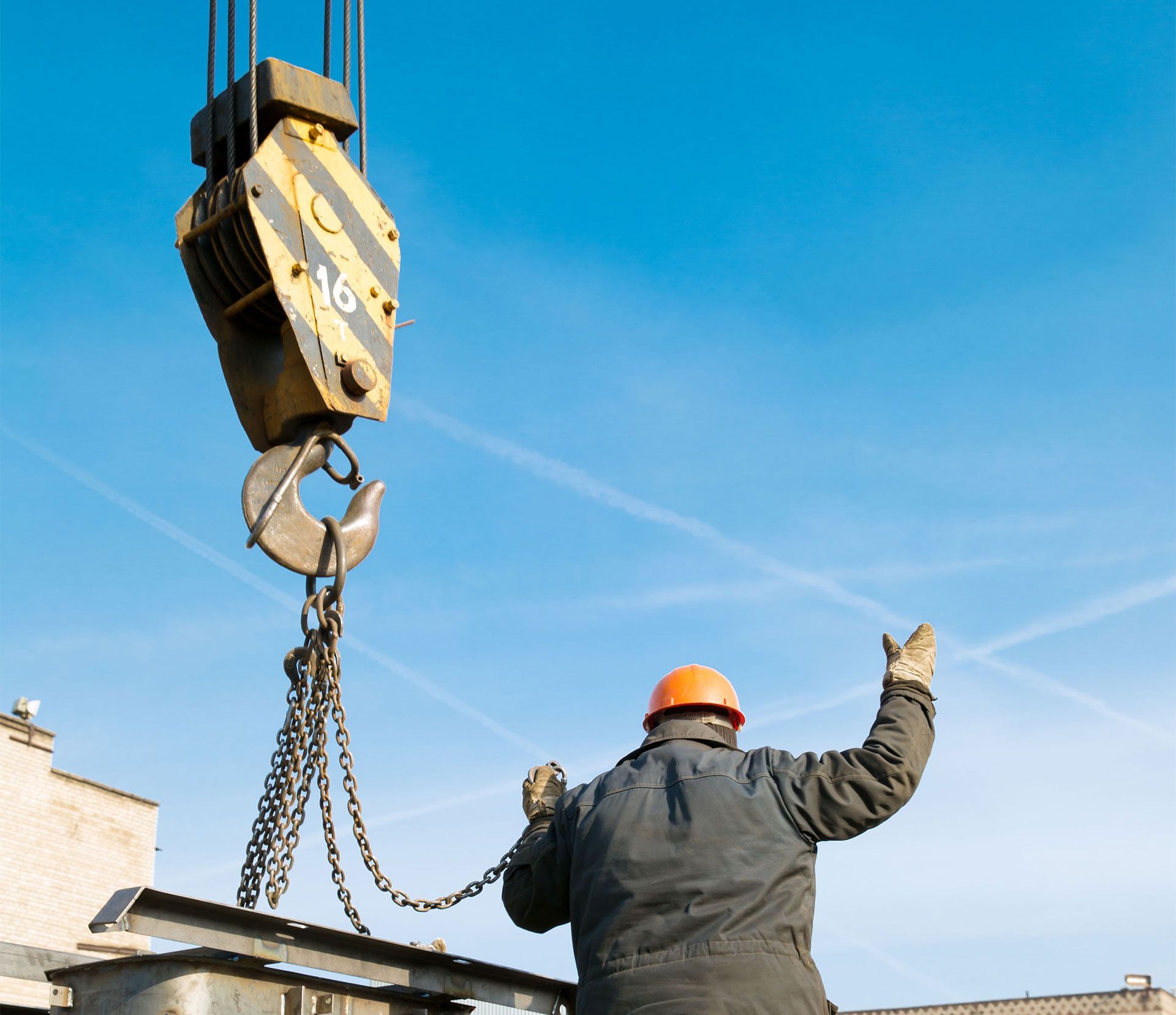 Construction worker guiding a crane hook with raised arms under a blue sky.