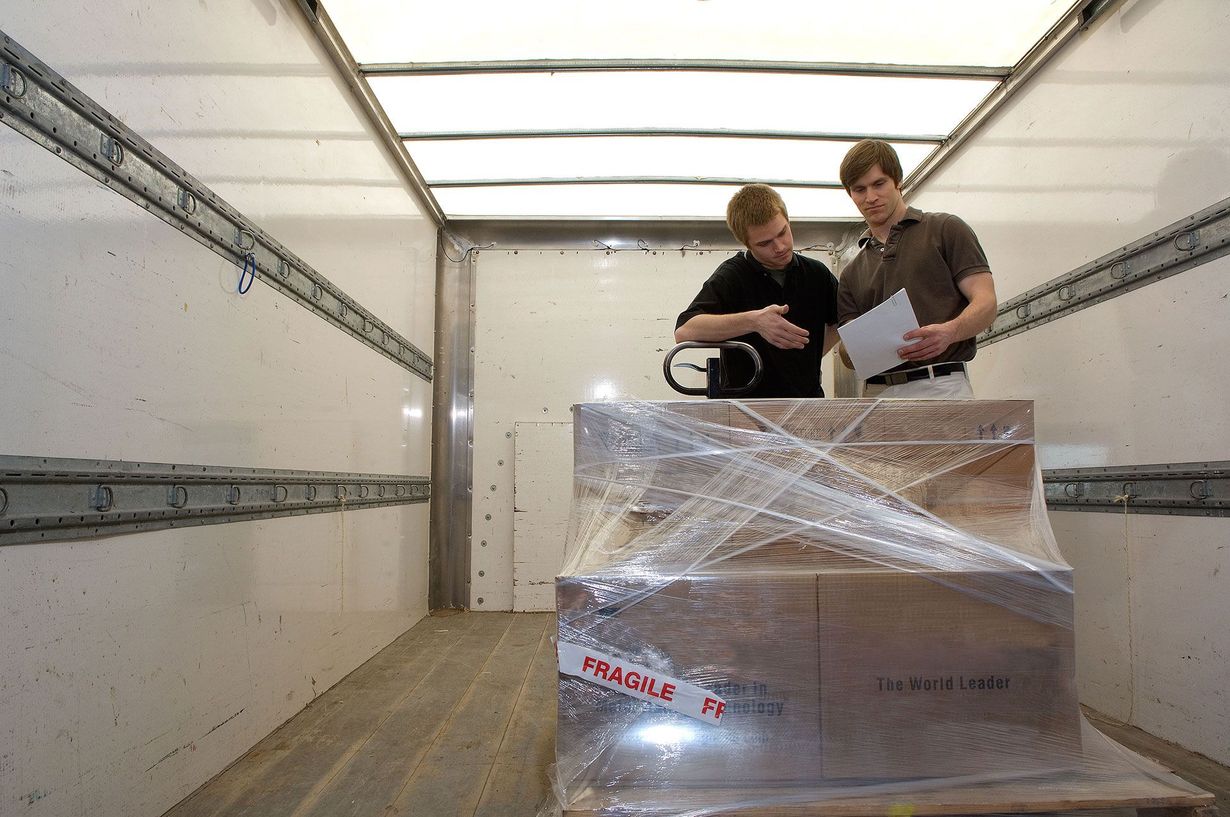Two men inspect a wrapped package inside a truck. One holds a document, the other handles a tool.