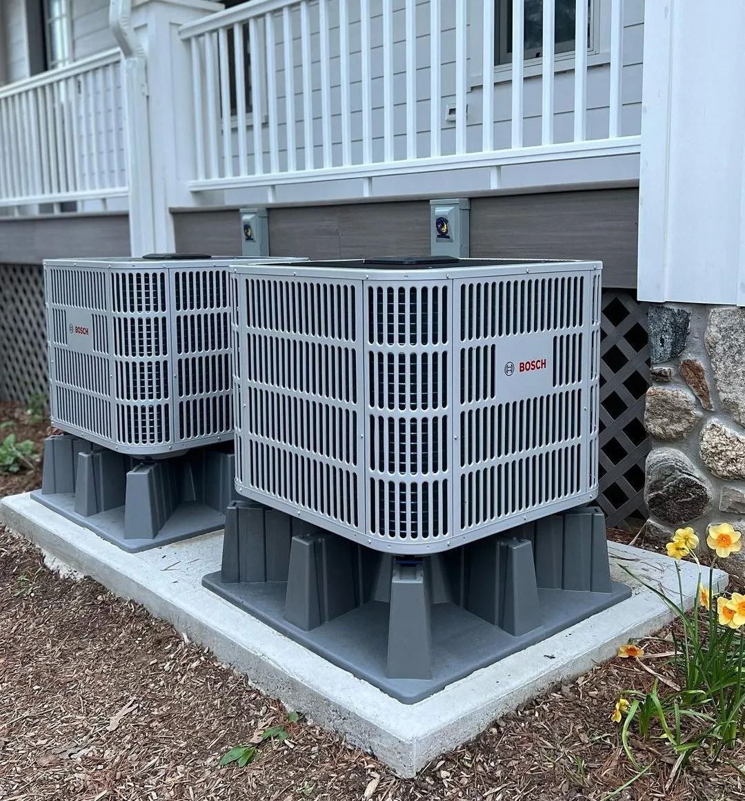 Two gray HVAC units sit on a concrete pad below a deck, near a stone foundation.