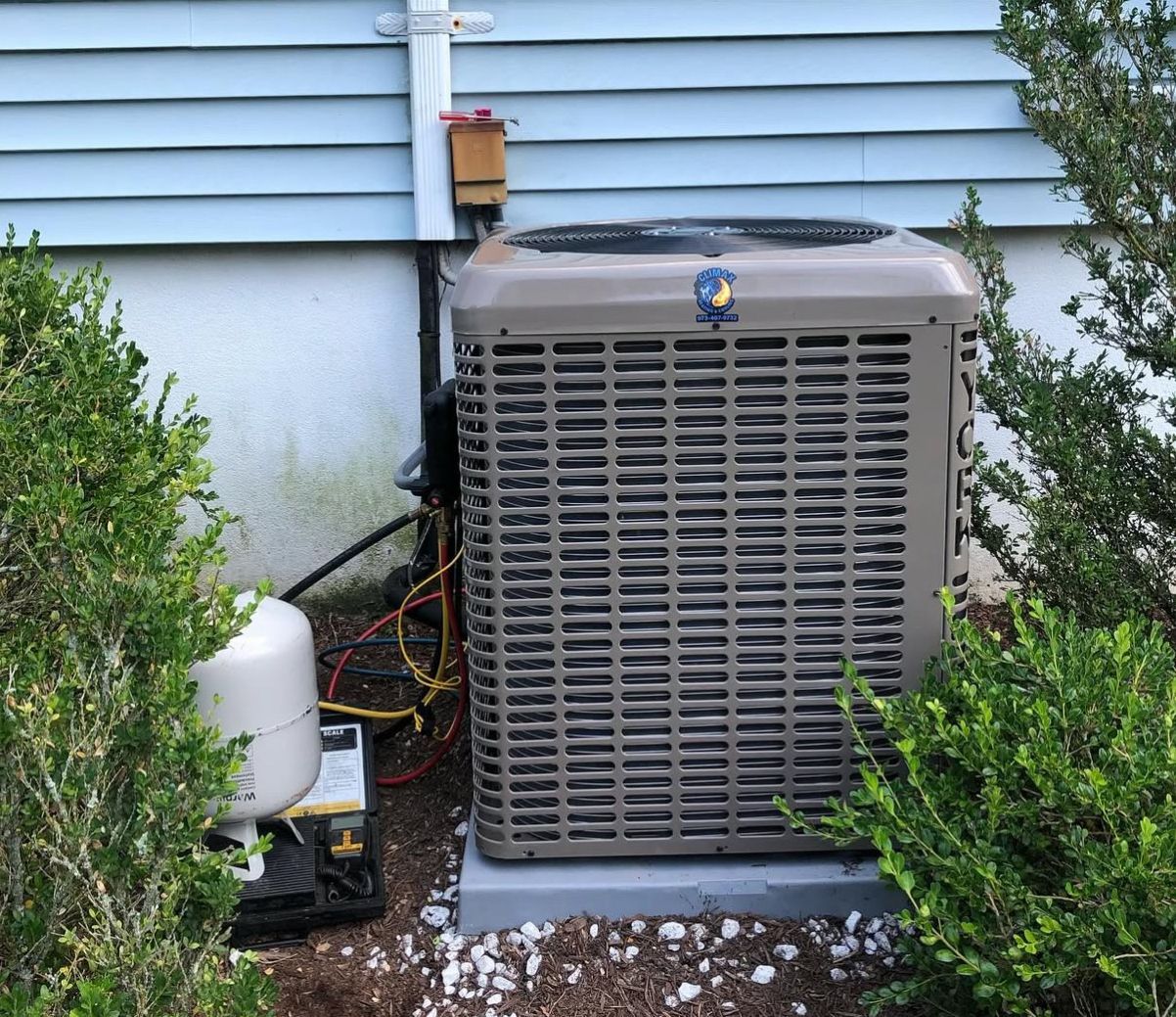 A beige outdoor HVAC condenser unit sits on a concrete pad against a light blue house exterior
