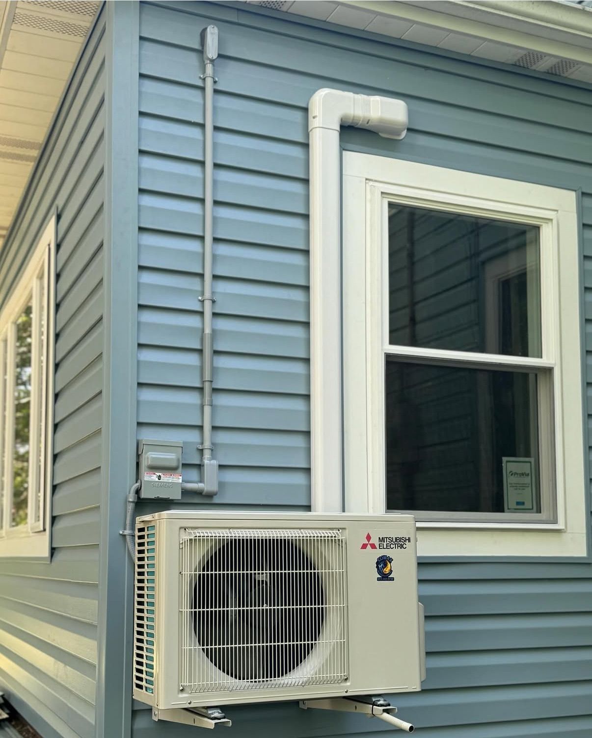 A Mitsubishi mini-split heat pump unit mounted on the exterior of a light blue vinyl-sided house 