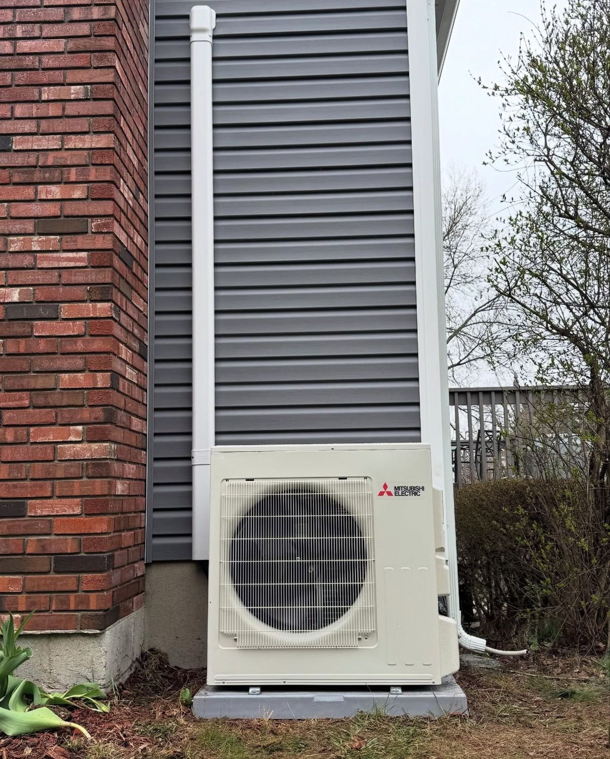 A Mitsubishi heat pump condenser  on a gray pad next to a brick wall and gray vinyl siding