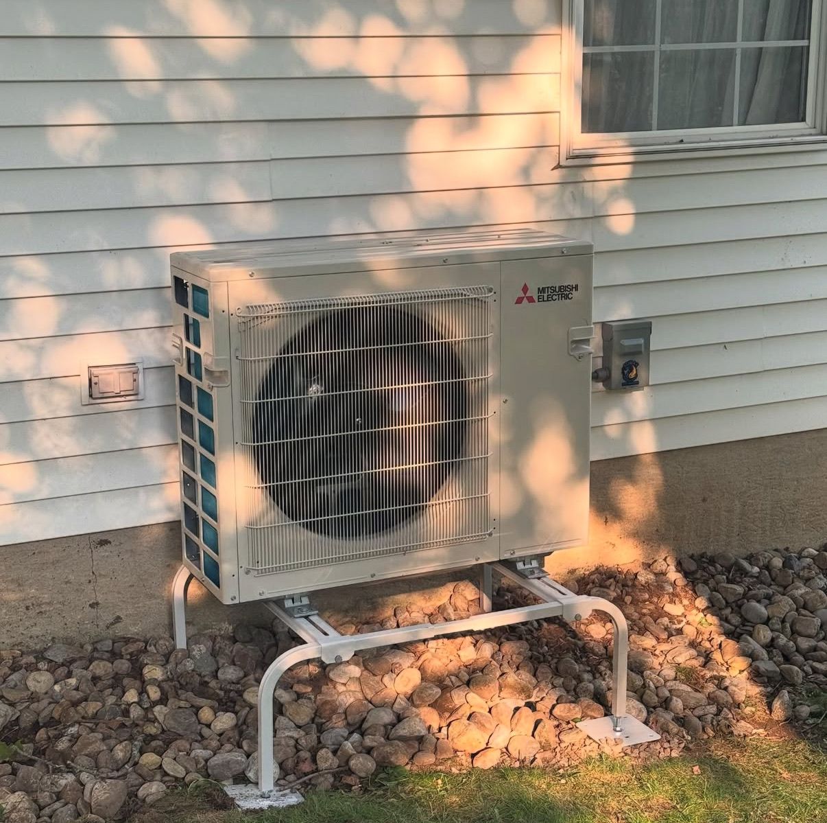 A Mitsubishi outdoor HVAC unit mounted on a metal stand above a gravel patch beside a white house wall