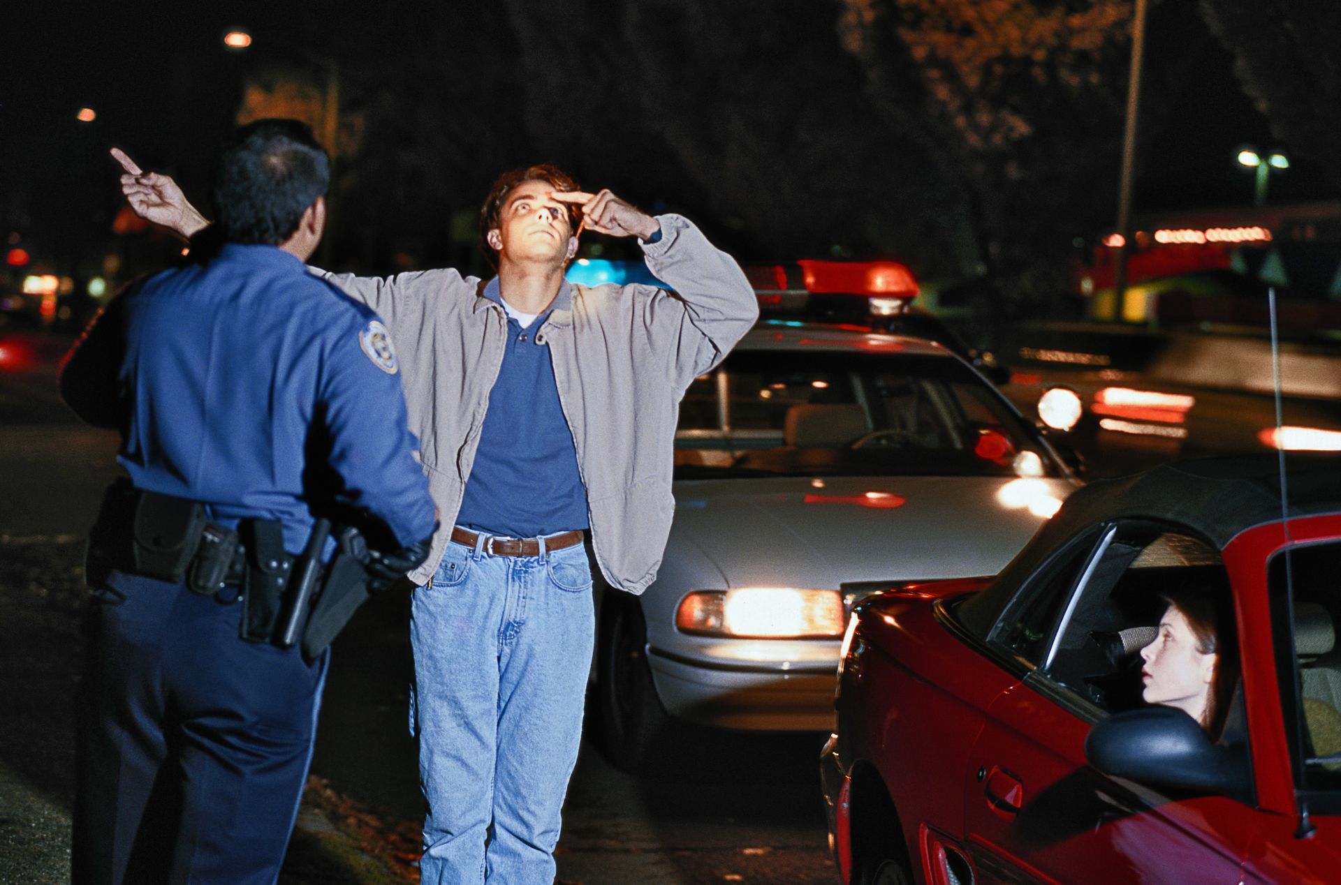 Police officer directing a driver, another in a red car, at night.