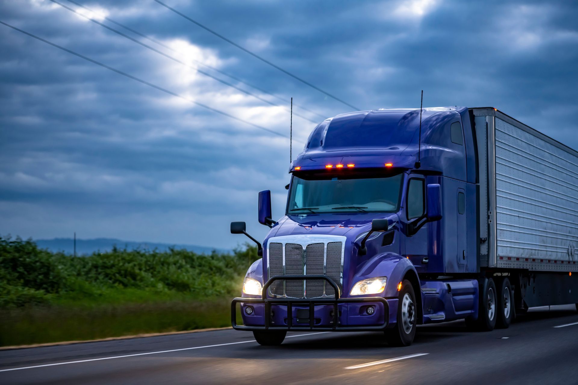 Blue semi-truck driving on a highway at dusk, carrying cargo.