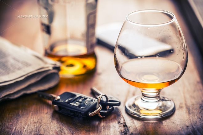 A brandy glass, a liquor bottle, and car keys on a wooden table next to a newspaper.