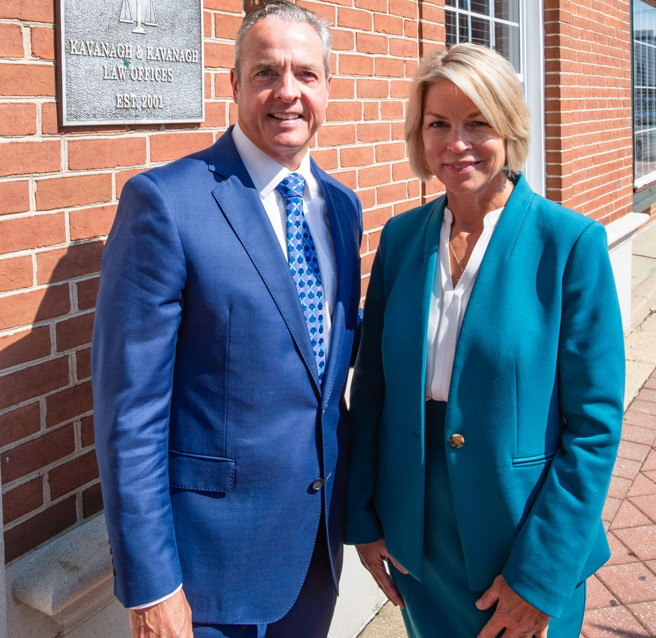 Man and woman stand outside a brick building. Man wears a blue suit, woman a teal suit.