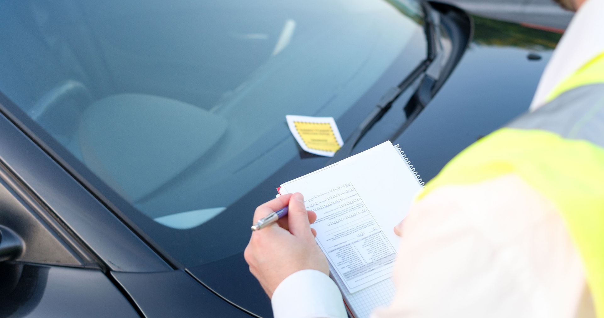 A parking enforcement officer in a yellow vest writes on a clipboard next to a car with a parking permit on the windshield.
