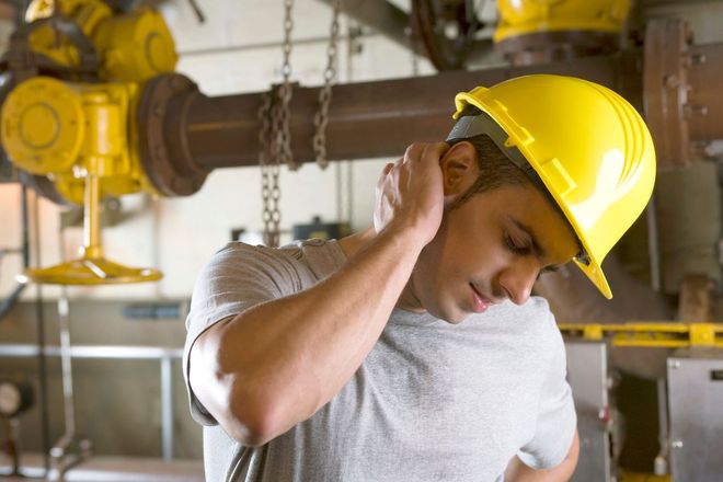 A construction worker in a yellow hard hat holding their neck in pain inside an industrial facility.