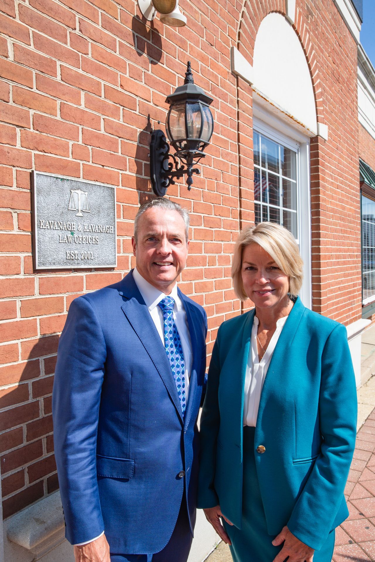Man and woman stand outside a brick building. Man wears a blue suit, woman a teal suit.