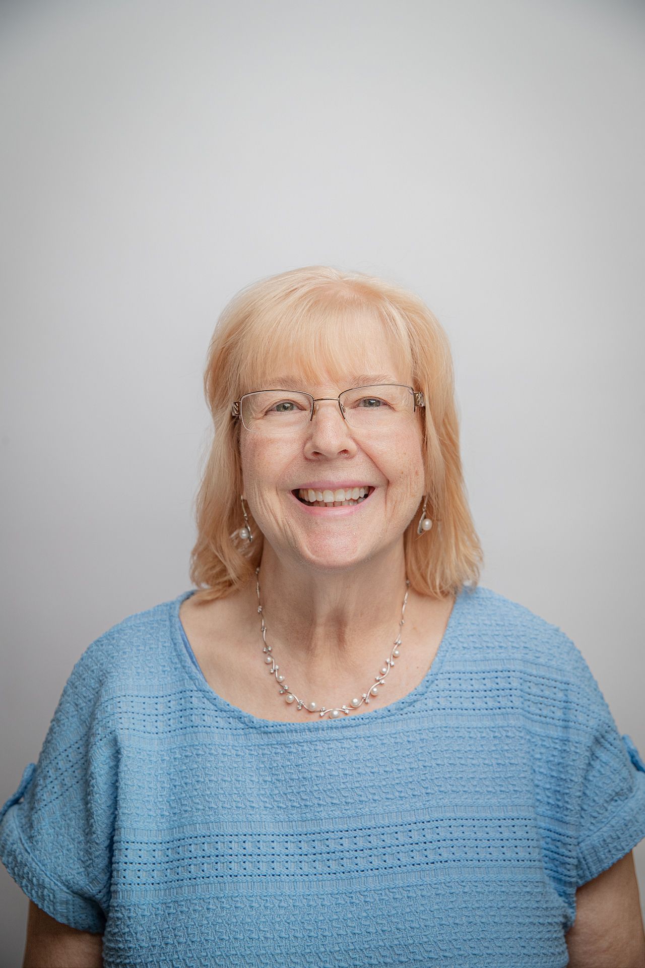 Woman with blonde hair and glasses smiles wearing a blue top, pearl necklace against a gray background.