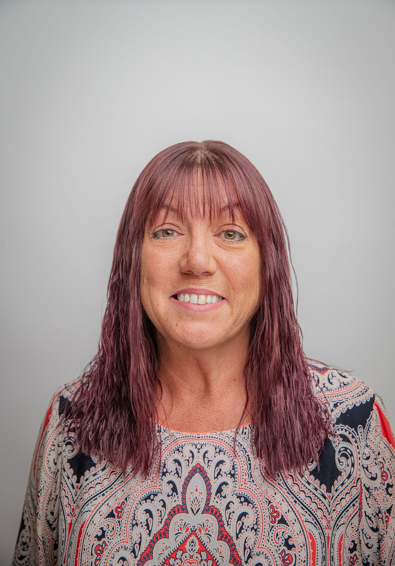 Woman with auburn hair and bangs, smiling, wearing a patterned blouse, against a white background.