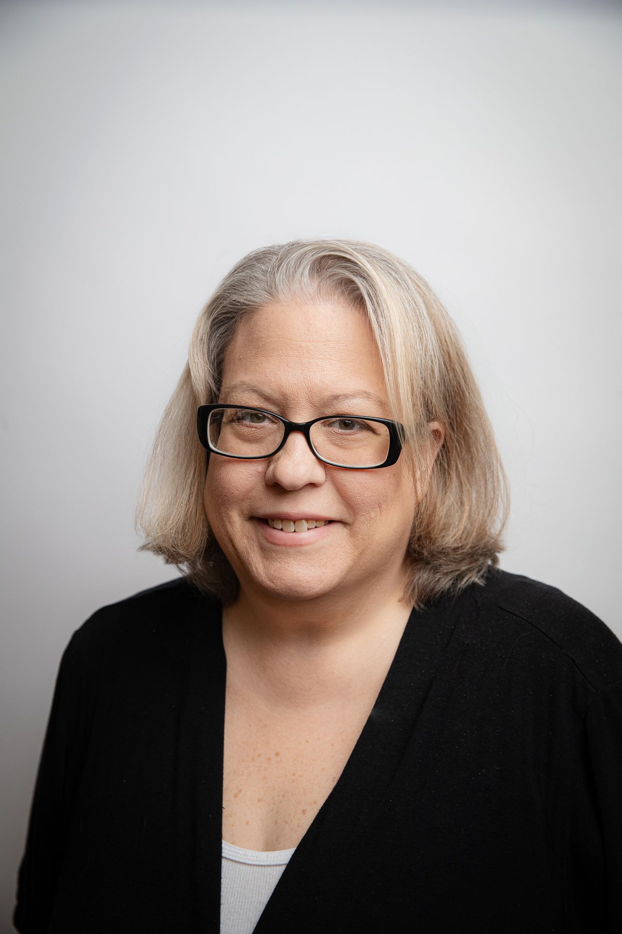 Woman wearing glasses and a black sweater smiles at the camera against a white background.