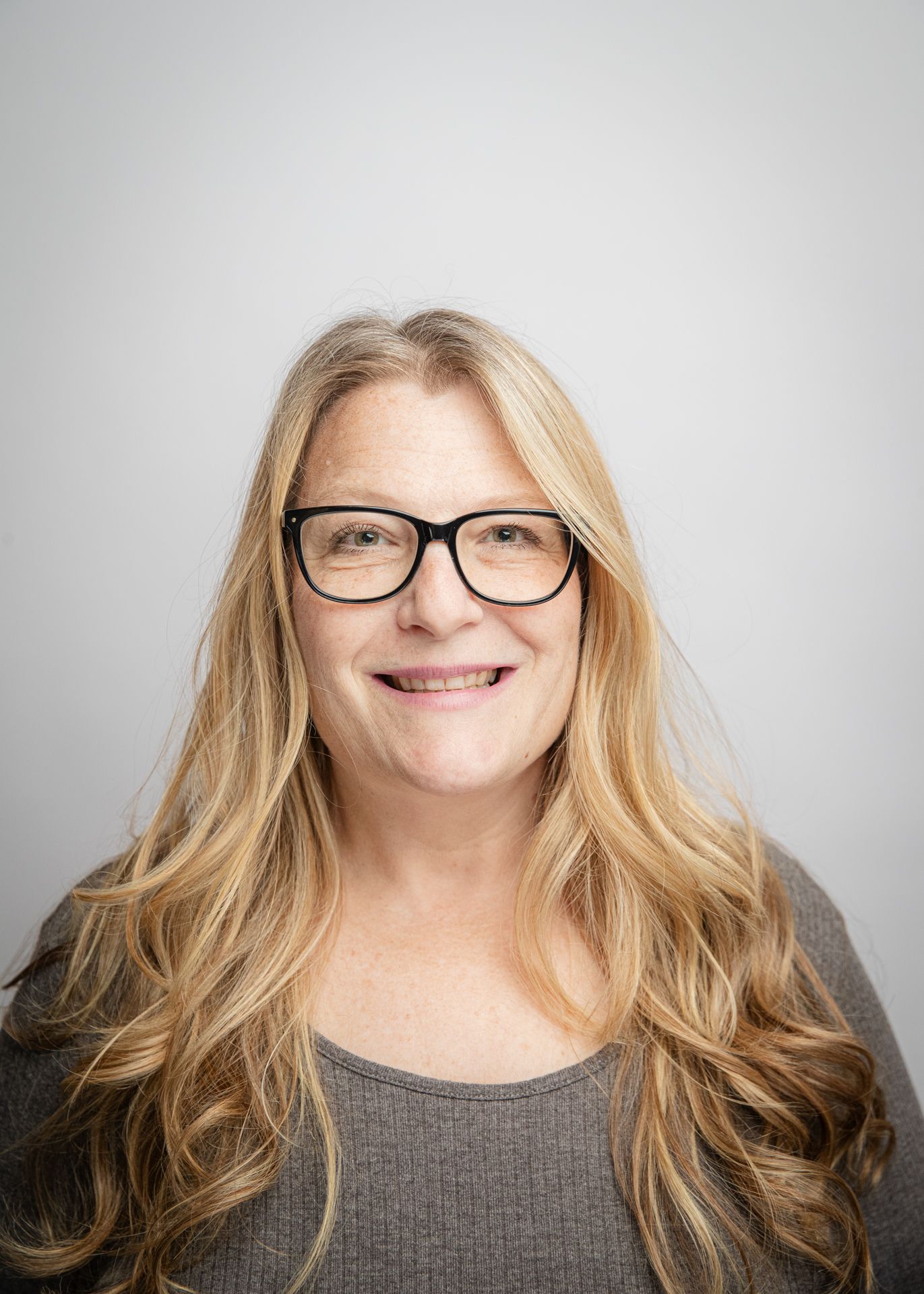 Woman with long blonde hair, wearing glasses and a gray sweater, smiling in front of a white background.