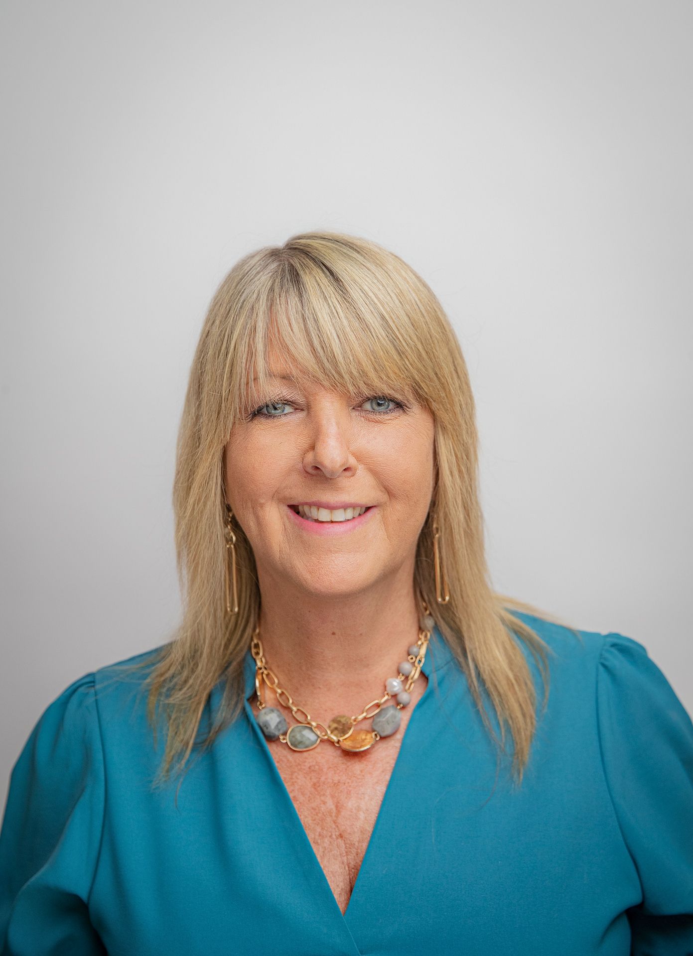 Woman with blonde hair wearing a teal blouse and beaded necklace, smiling at the camera.
