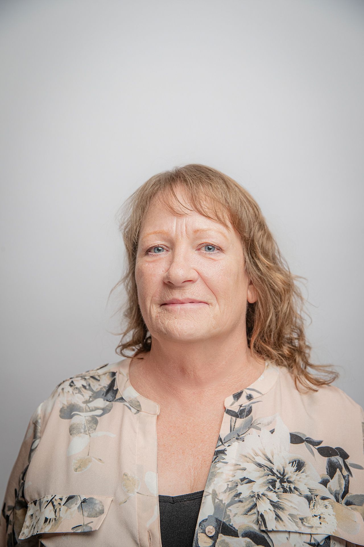 Woman with blonde, wavy hair smiles, wearing a floral-patterned blouse, against a plain background.