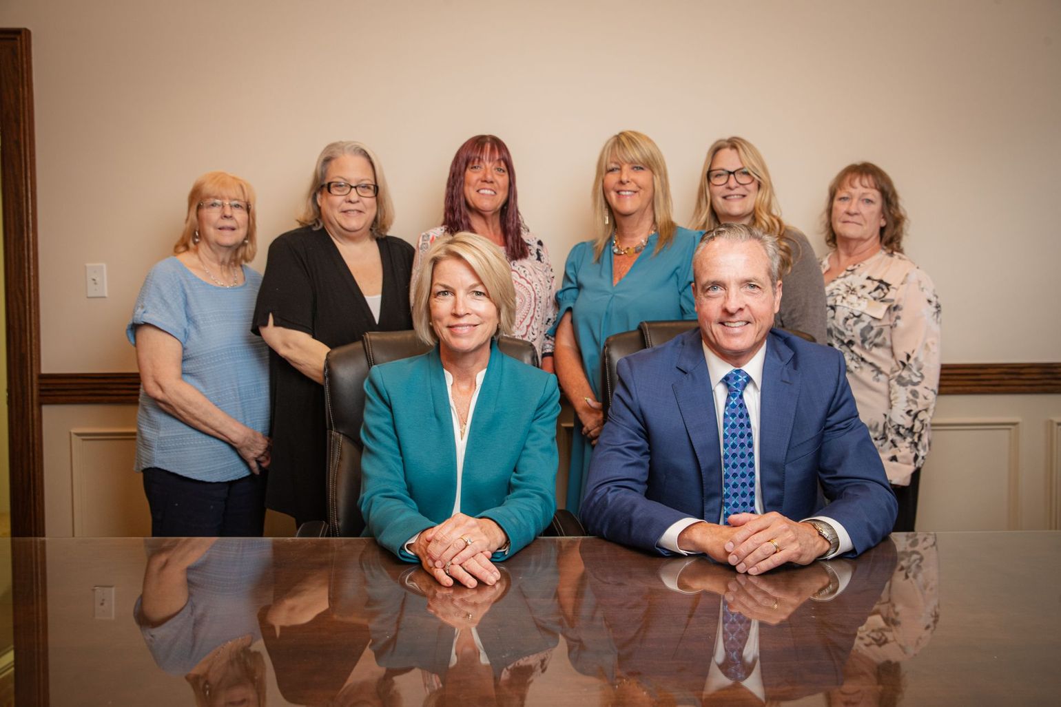 Business team seated and standing behind a conference table. People are wearing business attire, smiling.
