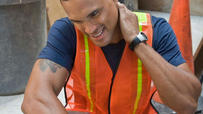 A construction worker in a yellow hard hat holding their neck in pain inside an industrial facility.