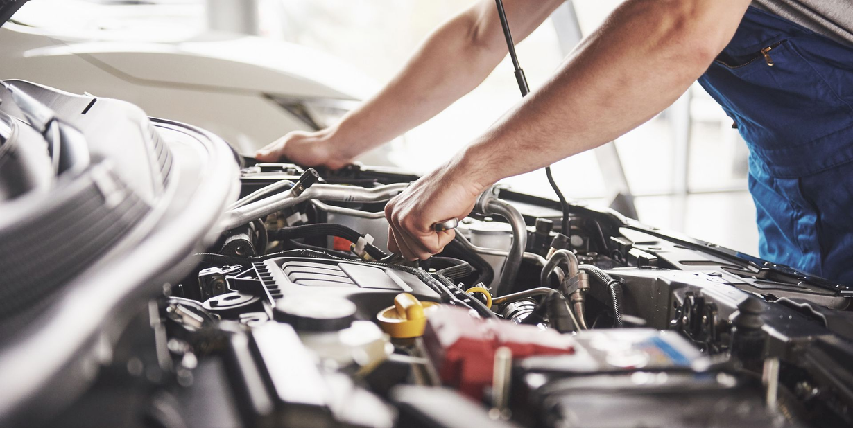 Mechanic working under the hood of a car in a garage, with tools and engine parts visible.
