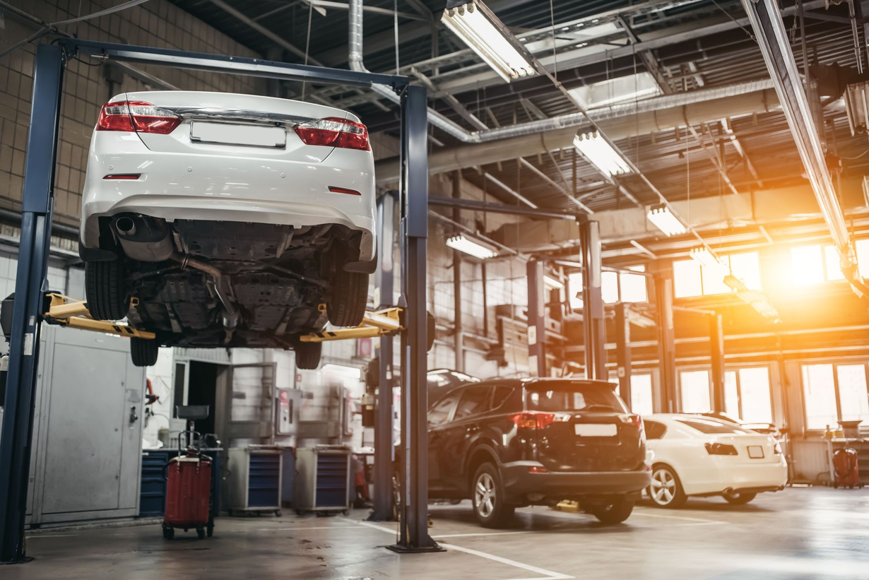Car lifted in auto repair shop with SUV below and sunlight streaming through the garage.