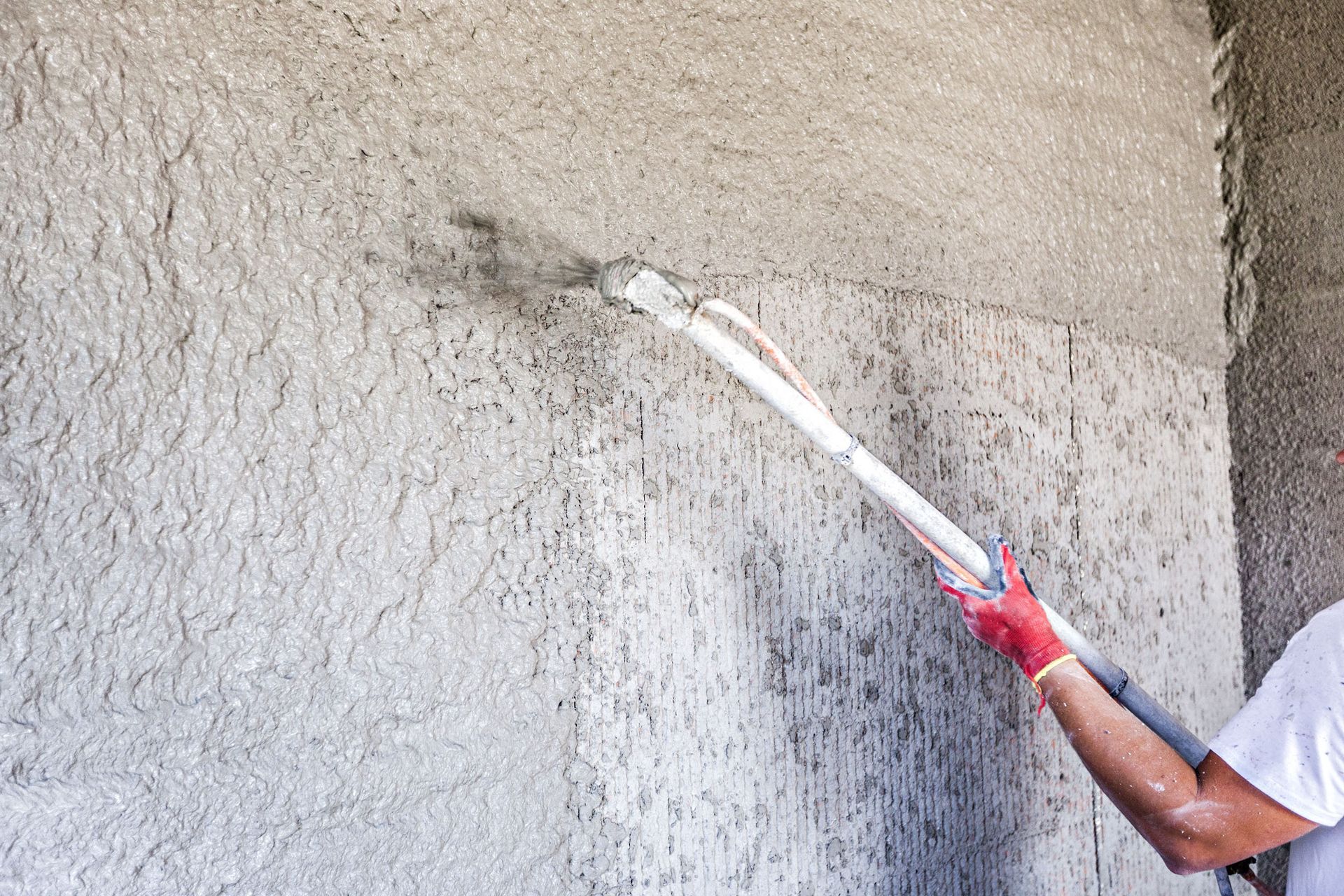 Person applying cement to wall with a spray gun. Wearing gloves and a white shirt.