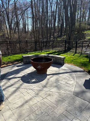 A patio with a fire pit, surrounded by a stone bench and a black fence, forest in the background.