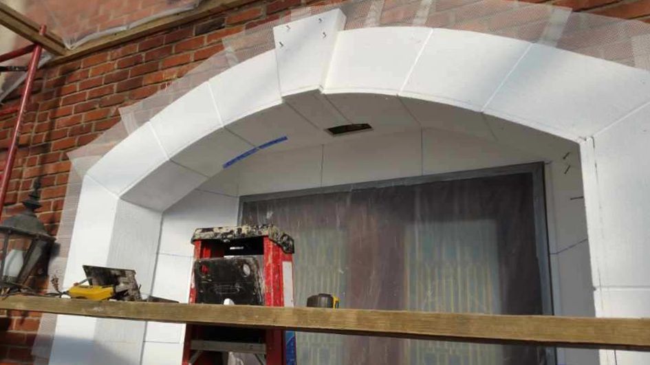 Arch doorway under construction, featuring white stone blocks. Red brick background, construction materials visible.