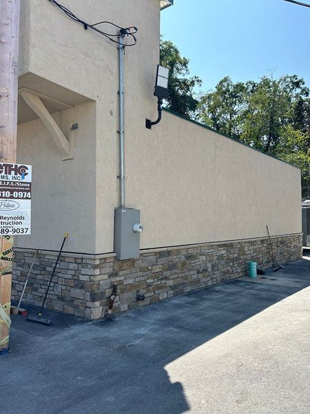 Exterior of building with light-colored stucco and stone veneer. Electrical conduit and panel visible. Blue sky overhead.