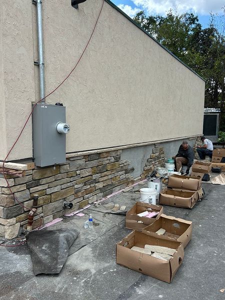 Stone veneer being installed on a building's exterior. Workers are applying mortar and placing stones.