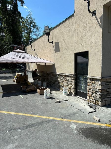 Exterior of a building under construction; stone work being added near door. Person sitting under an umbrella.
