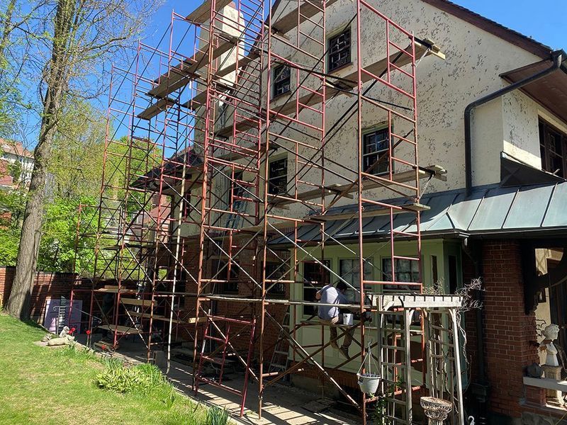 Red scaffolding surrounds a beige house undergoing renovations; a worker stands at the bottom.