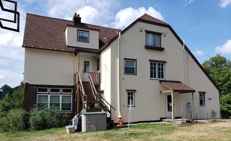 Beige stucco house with brown roof, windows, and wooden stairs. Bright blue sky visible.