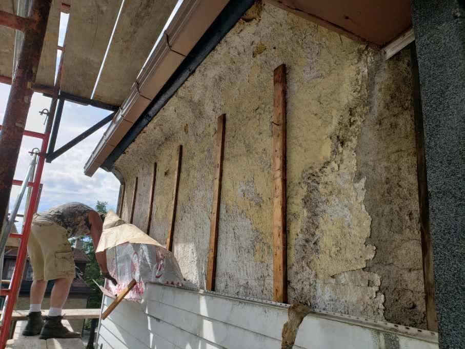 Man working on exterior wall with scaffolding. Peeling stucco, wooden supports, under eave.