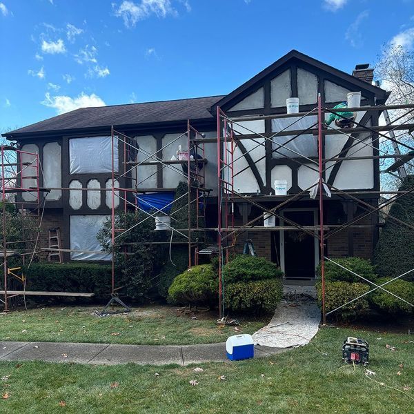House exterior under renovation; scaffolding and plastic sheeting cover areas. White paint visible, person on scaffolding.