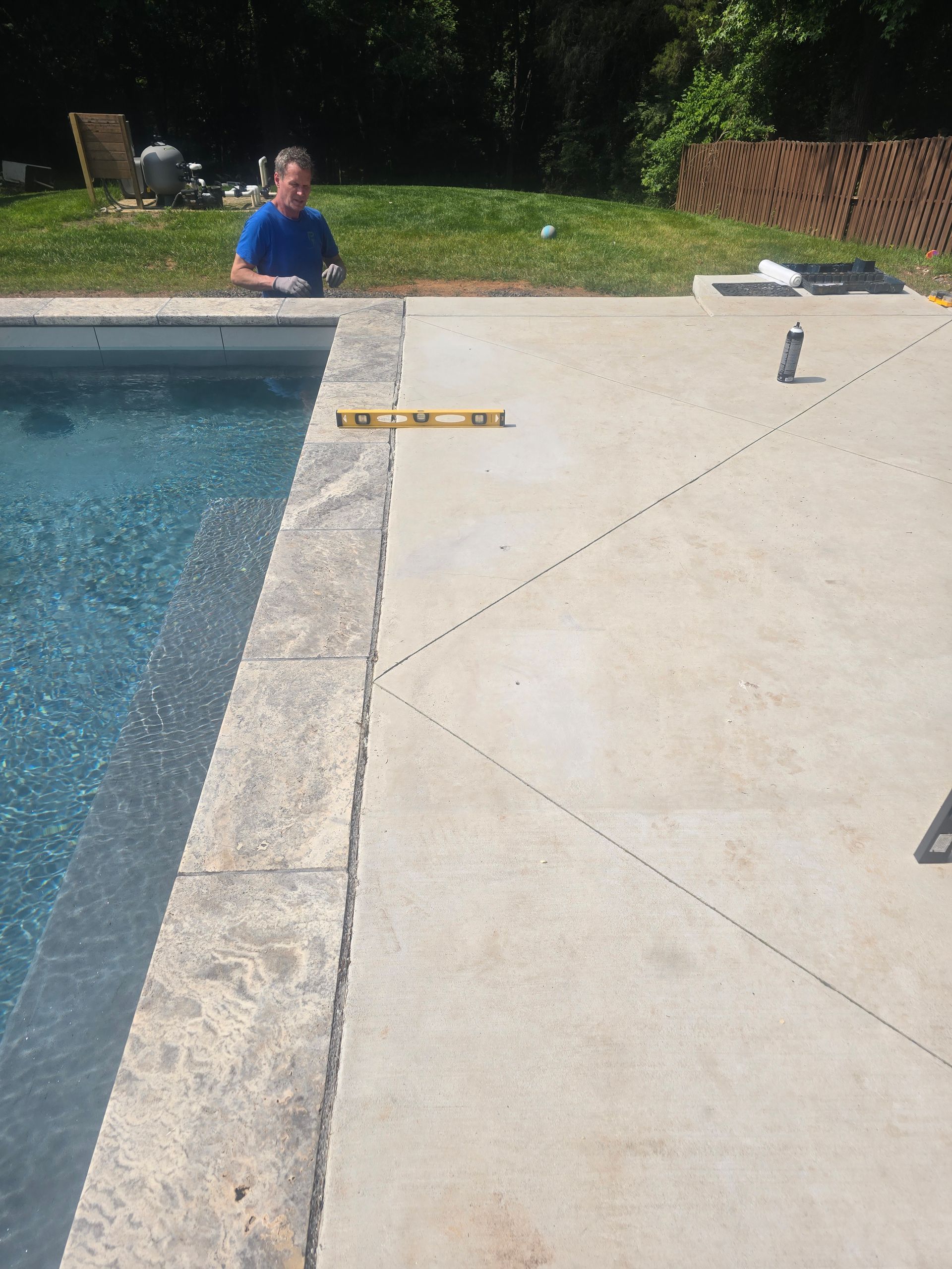 Man leveling concrete pool deck with blue water, green grass, and a fence in background.