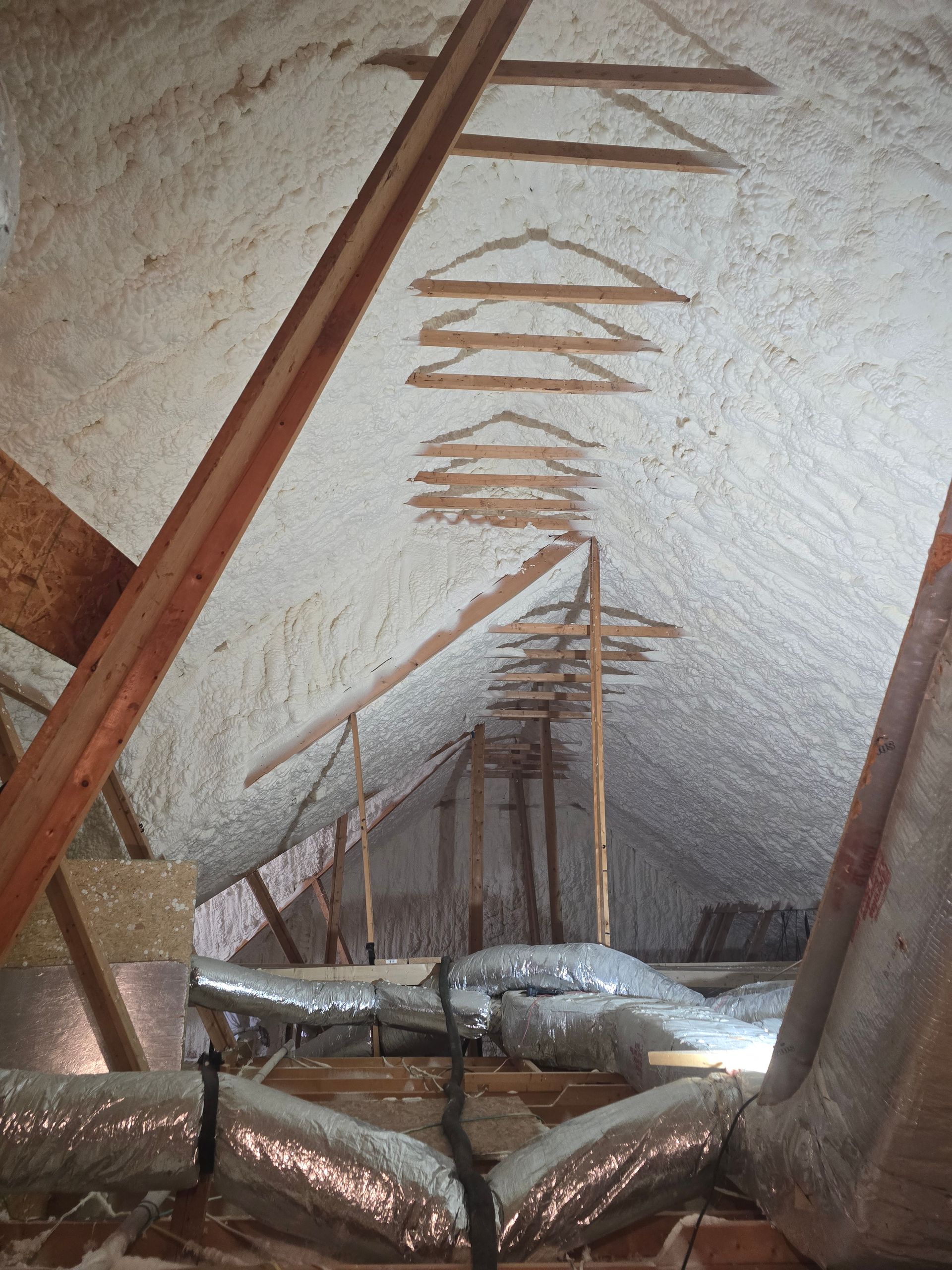Attic with spray foam insulation, ductwork, and wooden supports, seen from below.