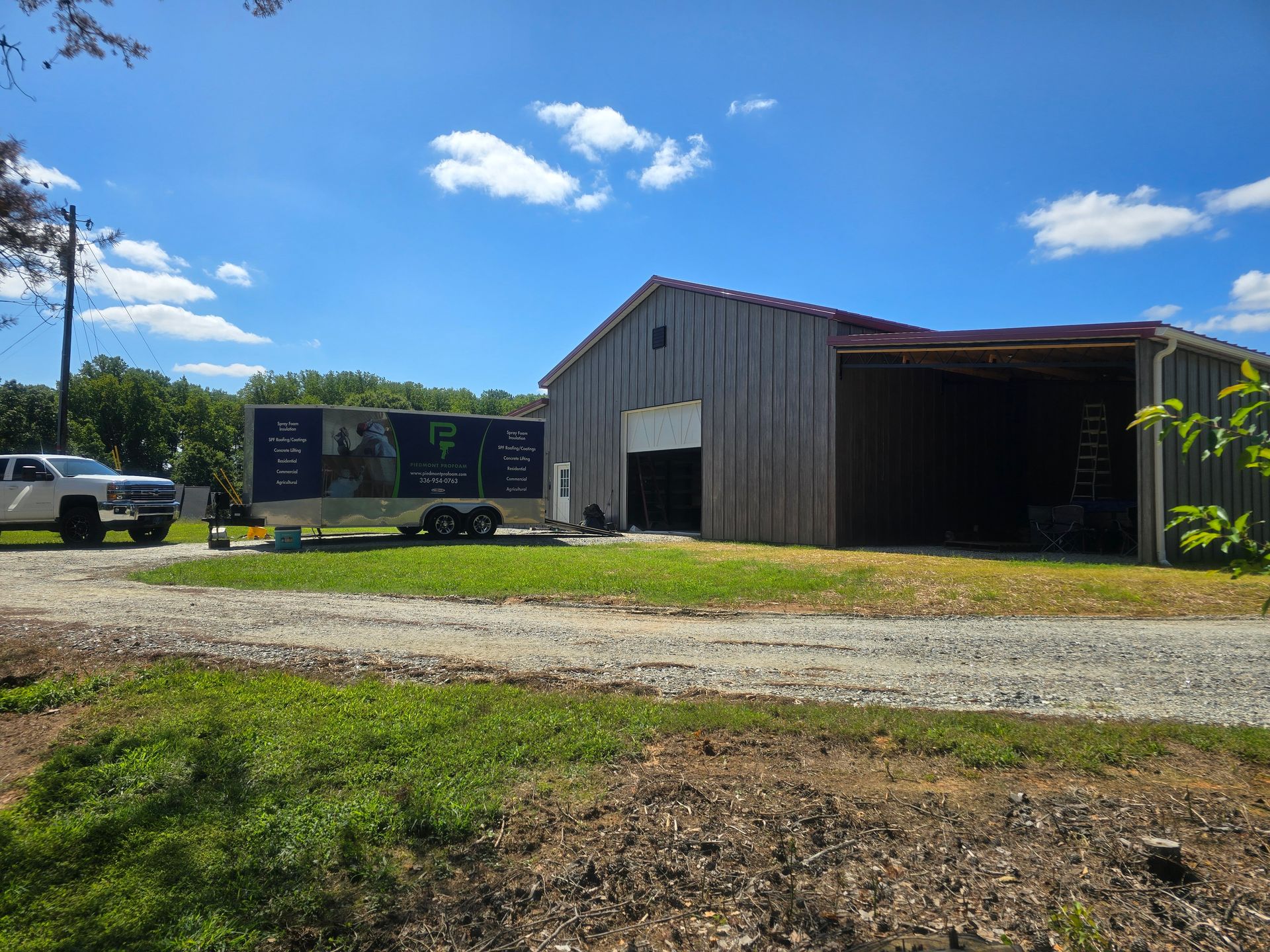 A white truck and trailer parked next to a weathered barn on a gravel driveway, under a sunny sky.