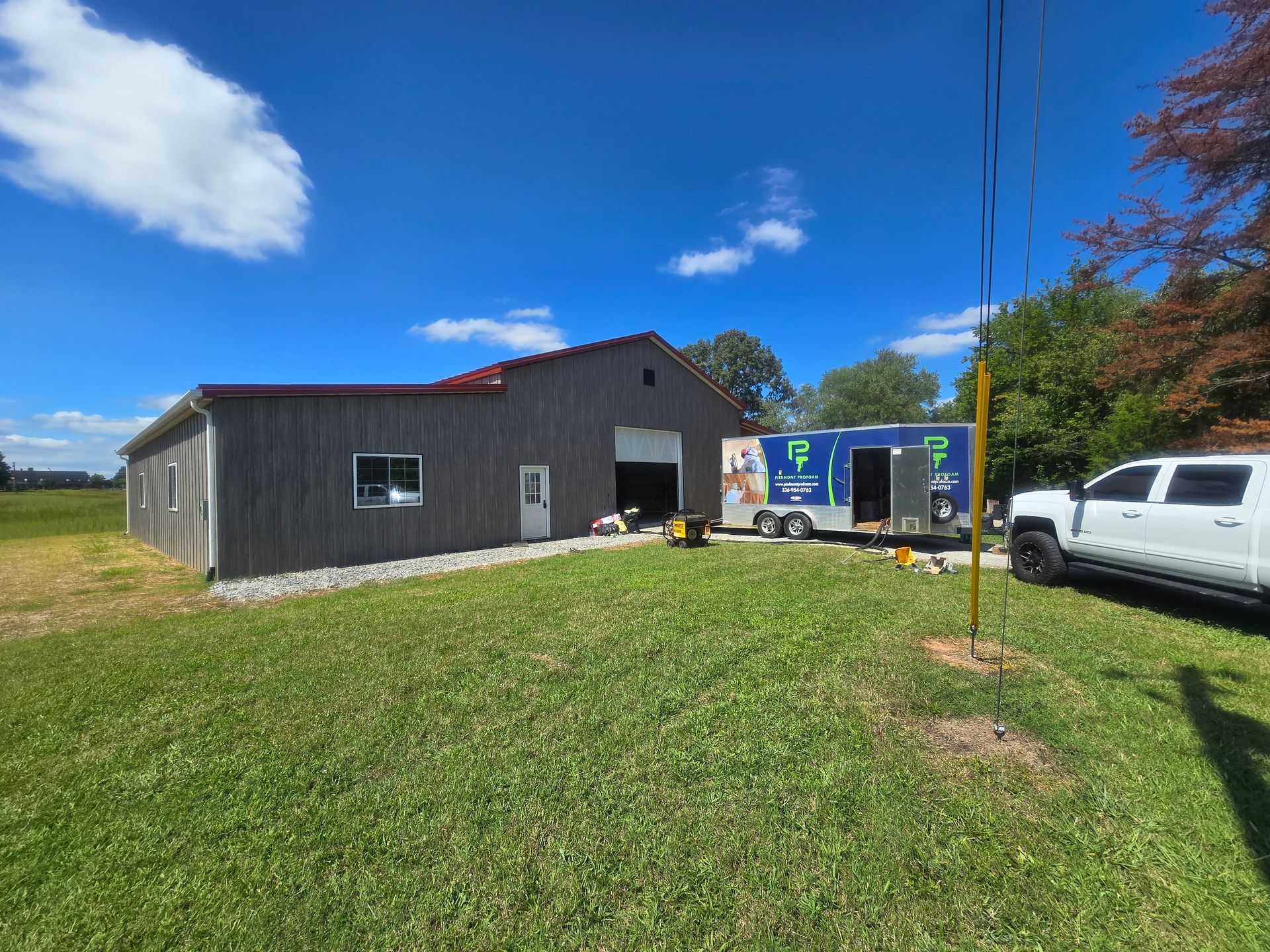 A gray barn with an open door, a mobile unit, and a white truck on a grassy field under a blue sky.