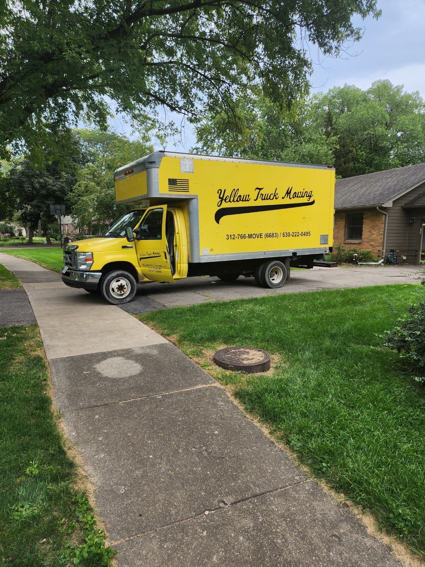 Yellow moving truck parked on a residential street. The truck is by a sidewalk and green grass.