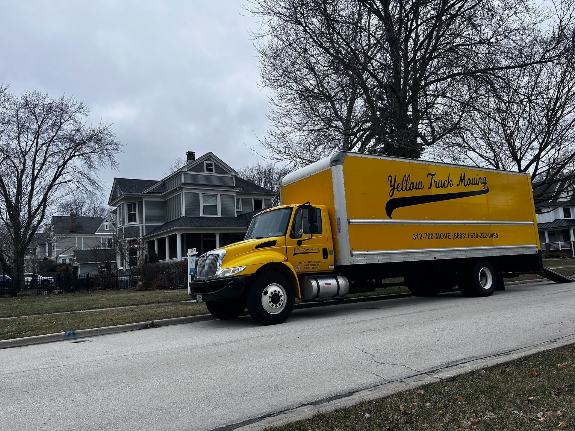 Yellow moving truck parked on a residential street in front of a gray house. Cloudy sky.