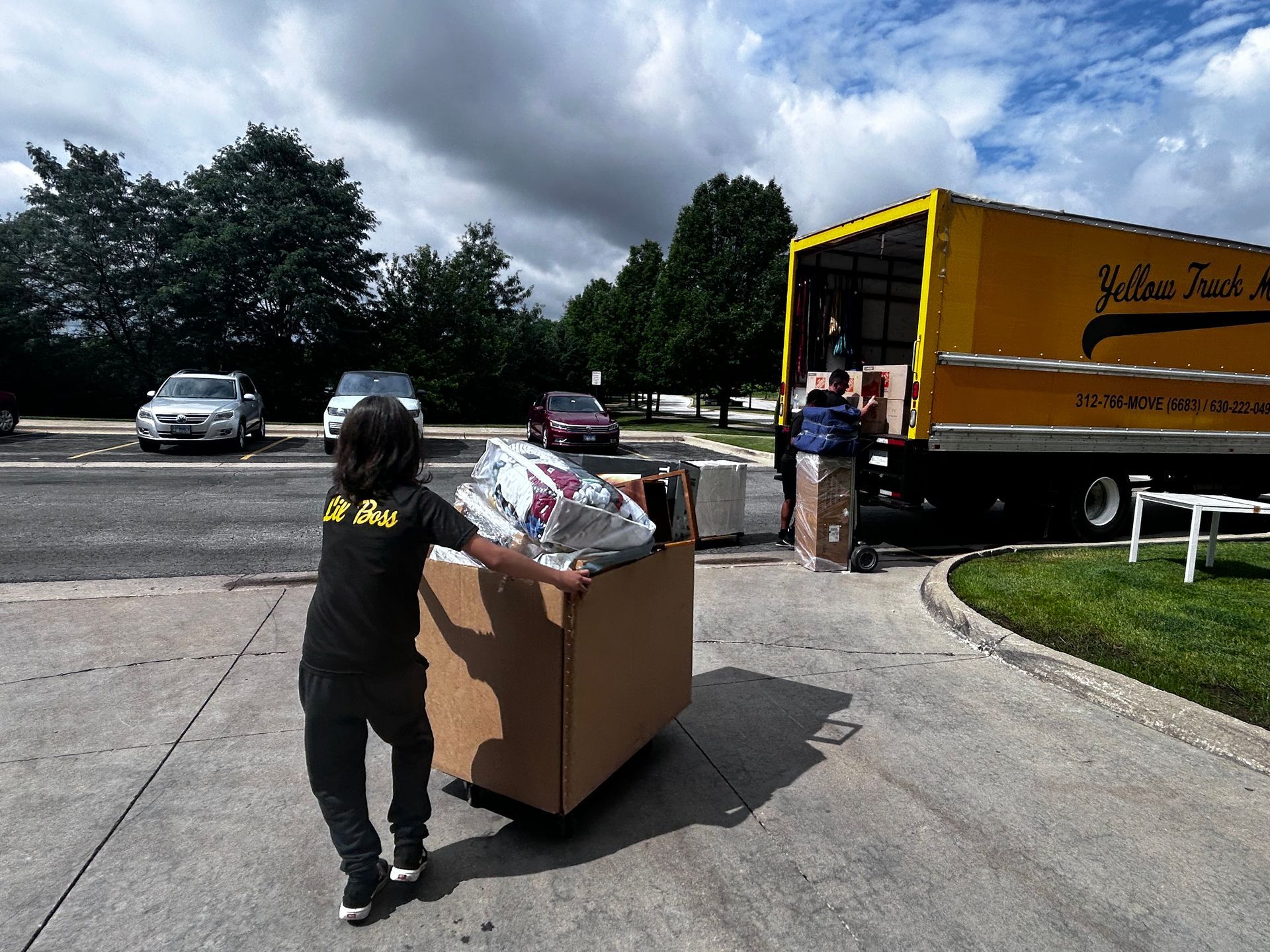 Person pushing a large box on a cart toward a yellow moving truck. Other people are near the truck.