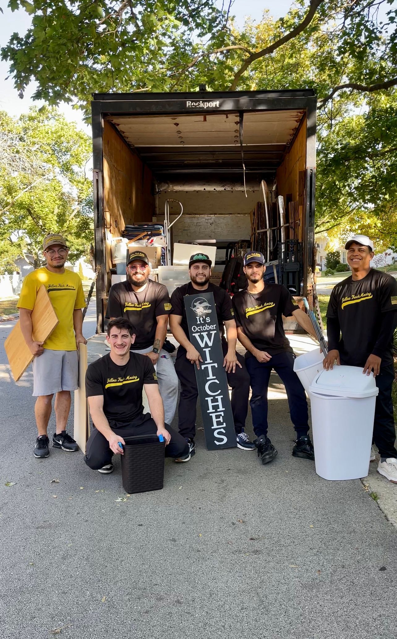 Movers stand in front of a truck, holding supplies. They wear black shirts, smiling.