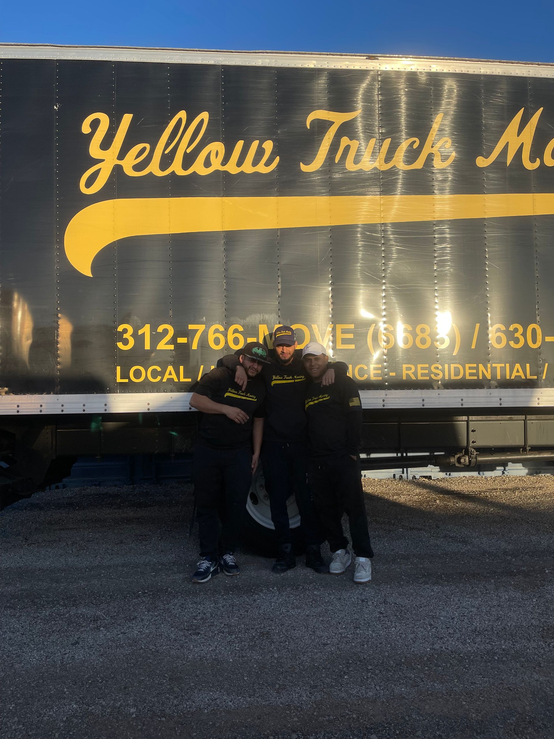 Three people in black uniforms stand in front of a Yellow Truck Moving truck.