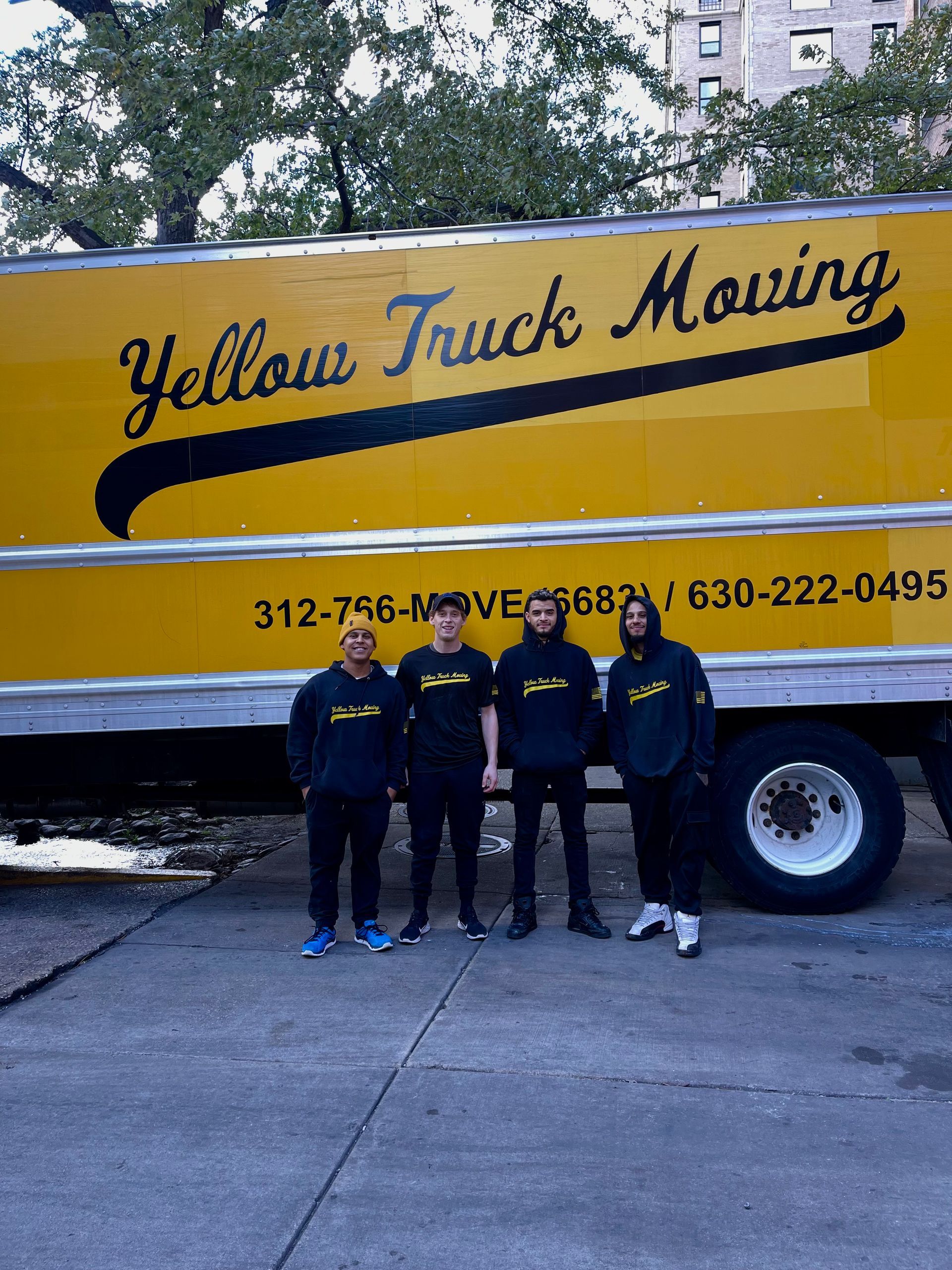 Four people in black hoodies stand in front of a yellow moving truck.